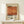 May include: Framed vintage-style art poster with orange and red abstract geometric composition, featuring "Mark Rothko" text and exhibition details. Shows cream-colored beige background with black typography. Displayed on light wood surface with small wrapped gift box beside frame. Warm natural lighting creates soft shadows on white wall background.