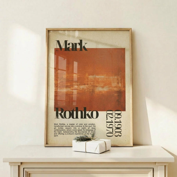May include: Framed vintage-style art poster with orange and red abstract geometric composition, featuring "Mark Rothko" text and exhibition details. Shows cream-colored beige background with black typography. Displayed on light wood surface with small wrapped gift box beside frame. Warm natural lighting creates soft shadows on white wall background.
