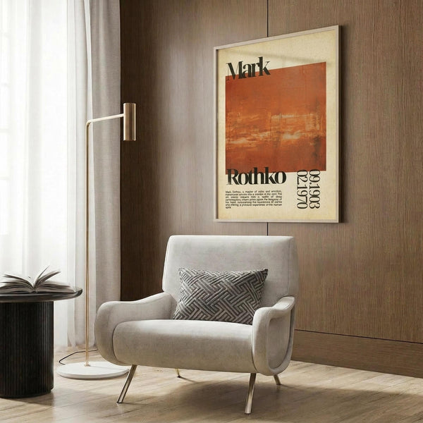 May include: Modern beige armchair with geometric patterned pillow in contemporary living room setting with wood paneled wall. Framed vintage-style art poster featuring orange and rust colored abstract painting with "Mark Rothko" text and dates "1903-1970" hangs above chair. Gold floor lamp with white marble base and open book on dark side table complete the styled interior scene.