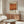 May include: Modern living room with white curved chair, glass oval coffee table, and wooden sideboard. Framed Mark Rothko exhibition poster with orange abstract artwork hangs on cream wall above furniture. Minimalist Scandinavian-style interior with natural wood tones and clean lines. Floor lamp and decorative objects complete the contemporary setting.