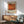 May include: Framed Mark Rothko exhibition poster with orange abstract artwork and black typography, displayed on white wall above wooden sideboard with table lamp. Mid-century modern living room setting features sage green upholstered armchair with wooden frame, round wooden coffee table, and natural sunlight casting geometric shadows across the scene.