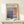 May include: Framed abstract art exhibition poster featuring Mark Rothko's signature color block style in soft blues, purples, oranges and grays. Wooden frame displays on white floating shelf against cream wall with small wrapped gift box below. Natural lighting creates gentle shadows.