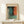 May include: Framed abstract art print in natural wood frame displaying soft color blocks in teal, orange, cream and dark tones in Rothko style, positioned on light wooden surface beside small white wrapped gift with evergreen sprig against cream colored wall