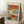 May include: Abstract art poster with soft geometric blocks in orange, blue, teal, yellow and white tones, displayed in light wood frame leaning against white wall. Scene shows cozy interior with coffee cup on wooden block side table, cream textured rug, and beige throw blanket. Warm natural lighting creates gentle shadows on the wall.