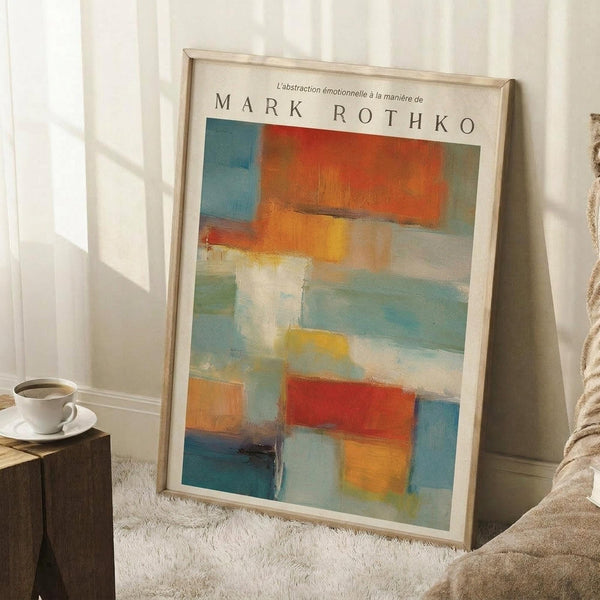 May include: Abstract art poster with soft geometric blocks in orange, blue, teal, yellow and white tones, displayed in light wood frame leaning against white wall. Scene shows cozy interior with coffee cup on wooden block side table, cream textured rug, and beige throw blanket. Warm natural lighting creates gentle shadows on the wall.