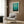 May include: Framed Mark Rothko art print with blue-green gradient color field displayed on wood paneled wall above cream upholstered armchair with geometric pillow. Modern floor lamp with brass finish and marble base stands nearby, with dark round side table holding open book. Neutral tones throughout contemporary living room setting with curtains and hardwood floors.