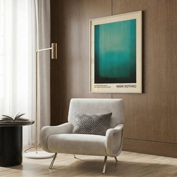 May include: Framed Mark Rothko art print with blue-green gradient color field displayed on wood paneled wall above cream upholstered armchair with geometric pillow. Modern floor lamp with brass finish and marble base stands nearby, with dark round side table holding open book. Neutral tones throughout contemporary living room setting with curtains and hardwood floors.