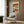 May include: Abstract colorful artwork with orange, blue, red, and white geometric blocks in white frame hanging on wood-paneled wall above beige upholstered armchair with geometric patterned pillow. Modern brass floor lamp and dark round side table with open book visible. Warm natural lighting from window with white curtains creates cozy reading nook atmosphere.