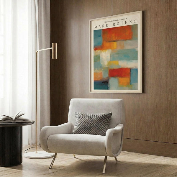 May include: Abstract colorful artwork with orange, blue, red, and white geometric blocks in white frame hanging on wood-paneled wall above beige upholstered armchair with geometric patterned pillow. Modern brass floor lamp and dark round side table with open book visible. Warm natural lighting from window with white curtains creates cozy reading nook atmosphere.