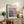 May include: Framed exhibition poster featuring Mark Rothko's abstract color field painting with soft blues, purples, and warm orange tones in geometric blocks. Displayed on rustic wooden sideboard alongside fern plant, ceramic vase, and books in cozy living room setting with beige armchair and neutral walls.