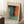 May include: Framed abstract art print leaning against white wall corner, featuring soft blended colors in teal, orange, and green with painterly brushstrokes. Cream-colored frame with white matting, positioned on textured white rug next to wooden side table with coffee cup and saucer, beige throw blanket visible at right edge.