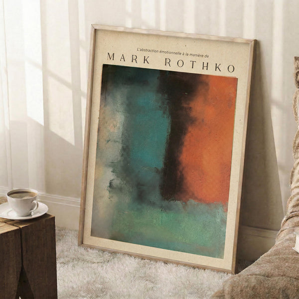 May include: Framed abstract art print leaning against white wall corner, featuring soft blended colors in teal, orange, and green with painterly brushstrokes. Cream-colored frame with white matting, positioned on textured white rug next to wooden side table with coffee cup and saucer, beige throw blanket visible at right edge.