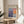 May include: Framed exhibition poster for Mark Rothko displayed on round wooden side table next to beige upholstered chair. Poster features abstract color blocks in blues, purples, oranges and whites with gallery text and dates. Natural lighting creates soft shadows across cream colored wall. Green plant leaves visible in corner.