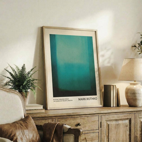 May include: Framed Mark Rothko museum poster featuring gradient blue and turquoise color blocks, displayed on rustic wooden sideboard in warm-toned living room with fern plant, ceramic vase, books, and cream upholstered chair with brown leather throw pillow