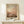 May include: Abstract expressionist painting print in warm earth tones featuring soft brushstrokes in peachy pink, golden yellow, and brown hues that blend into cloudy, atmospheric forms. The artwork is displayed in a light wooden frame with white matting, positioned on a pale wooden console table. A small white gift box with natural twine and evergreen sprig sits in front of the frame against a cream-colored wall.