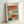 May include: Abstract expressionist art print in light wooden frame leaning against white wall. Features soft rectangular blocks of color in orange, red, blue, turquoise, yellow and white with blended, painterly edges. French text at top reads "L'abstraction émotionnelle à la manière de" followed by "MARK ROTHKO" in bold letters. Warm natural lighting creates subtle shadows on white surface.