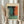 May include: Person in beige clothing holding framed abstract artwork with teal, orange, and dark color blocks in Rothko style. Light wooden frame against cream backdrop with dried pampas grass visible on left side. Soft natural lighting creates gentle shadows.