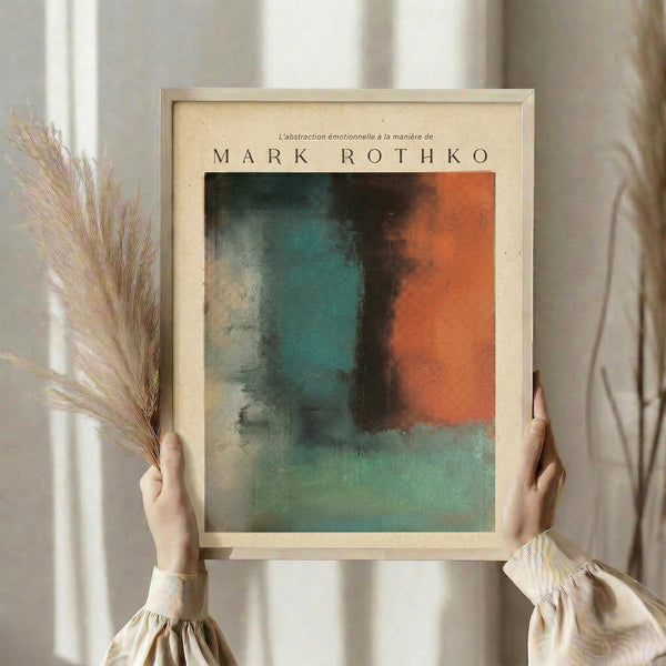 May include: Person in beige clothing holding framed abstract artwork with teal, orange, and dark color blocks in Rothko style. Light wooden frame against cream backdrop with dried pampas grass visible on left side. Soft natural lighting creates gentle shadows.
