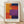 May include: Framed abstract art print with bold rectangular color blocks in orange, yellow, red, navy blue, and purple, displayed in light wood frame with white matting against white textured wall on wooden floor with natural window light creating soft shadows