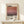 May include: Framed art poster featuring Mark Rothko's name in bold black letters above horizontal bands of warm gradient colors transitioning from cream to coral to deep burgundy, with "Rothko" and "1903-1970" text below. Light wood frame displayed on white wooden surface against cream wall, with small wrapped gift box nearby.