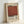 May include: Framed vintage-style exhibition poster featuring a large rust-red square on cream background with "Mark Rothko" text and dates "02.19.70 - 05.03.69". Light wooden frame leans against white wall, showing aged paper texture and small descriptive text about the artist's color mastery.