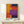 May include: Colorful abstract art poster with geometric blocks in orange, yellow, red, navy blue, and purple tones displayed in thin wooden frame on white wooden shelf. Small wrapped gift box with evergreen sprig sits beside the framed artwork against cream colored wall with natural lighting creating soft shadows.