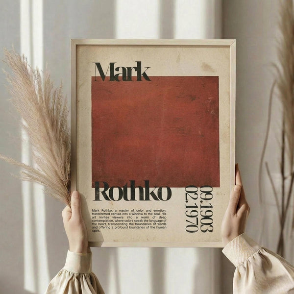 May include: Hands holding framed art print featuring rust-colored rectangular block with "Mark Rothko" text and biographical description below, styled as vintage exhibition poster. Natural wood frame against cream textured background with dried pampas grass visible on left side. Person wearing beige linen clothing with gathered cuff sleeves.