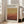 May include: Framed exhibition poster leaning against beige armchair in bright living room with large windows. Poster features warm gradient from golden yellow to deep rust red with black text header and exhibition details. Light wood frame, cream matting. Natural sunlight creates soft shadows on white walls and light hardwood floors.