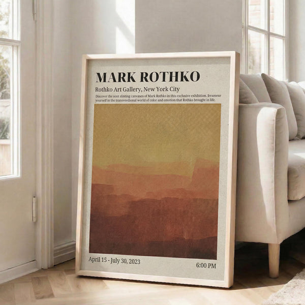 May include: Framed exhibition poster leaning against beige armchair in bright living room with large windows. Poster features warm gradient from golden yellow to deep rust red with black text header and exhibition details. Light wood frame, cream matting. Natural sunlight creates soft shadows on white walls and light hardwood floors.
