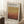 May include: Framed art poster leaning against white wall corner featuring abstract landscape in warm gradient tones from golden yellow to deep rust red. Light wood frame, displayed on white textured rug beside wooden side table with white coffee cup and saucer. Natural window light creates soft shadows on wall.