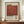 May include: Framed vintage-style poster with cream background and large rust-red square, featuring "Mark Rothko" in bold black text and biographical description below. Wooden frame sits on mid-century modern credenza beside white ceramic vase and stacked books against cream paneled wall.