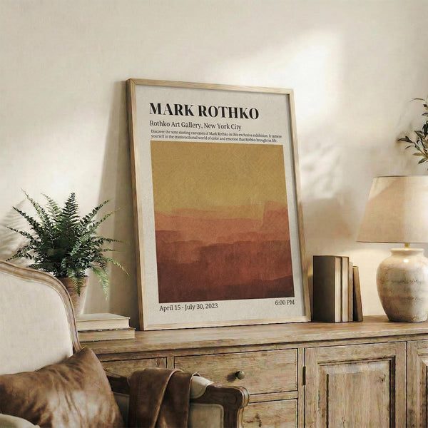 May include: Framed exhibition poster with Mark Rothko text and warm gradient artwork in golden yellow to deep rust tones, displayed on rustic wooden sideboard alongside fern plant, ceramic vases, and stacked books in cozy living room setting with beige chair and soft natural lighting