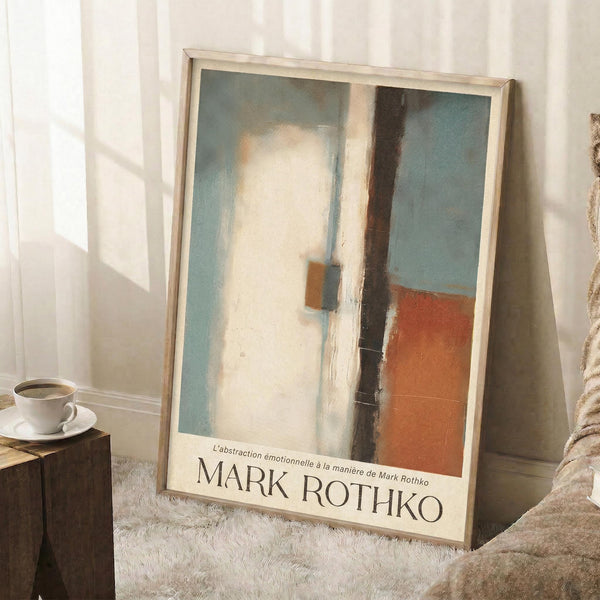 May include: Framed abstract art print leaning against white wall in cozy room setting. The artwork features soft geometric shapes in muted blue, cream, orange and brown tones with painterly brushstrokes. Light wood frame sits on white textured rug next to wooden side table with white coffee cup. Natural sunlight creates gentle shadows on the wall.