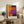 May include: Framed abstract art print with bold geometric color blocks in orange, red, purple, and navy blue, displayed on rustic wooden sideboard alongside potted fern, ceramic vase, and books in cozy living room setting with beige upholstered chair and neutral walls