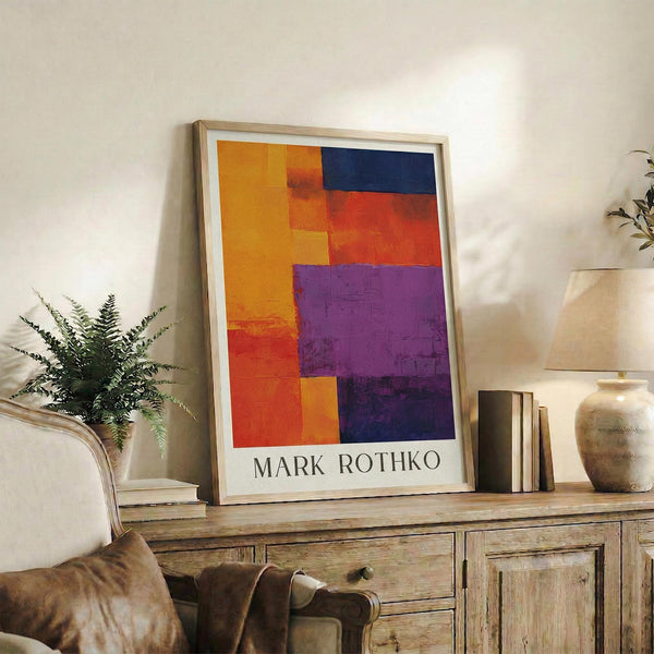 May include: Framed abstract art print with bold geometric color blocks in orange, red, purple, and navy blue, displayed on rustic wooden sideboard alongside potted fern, ceramic vase, and books in cozy living room setting with beige upholstered chair and neutral walls