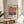 May include: Framed art print featuring Mark Rothko's name and exhibition details with a gradient abstract painting in warm coral, orange, and purple tones. The poster is displayed on a round wooden stool next to a beige upholstered chair with plant shadows cast across white walls in natural sunlight.