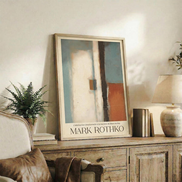 May include: Abstract art print with soft blue, white, orange and dark vertical forms in light wood frame, displayed on rustic wooden console table alongside fern plant, ceramic vase, stacked books and leather armchair in warm neutral living room setting