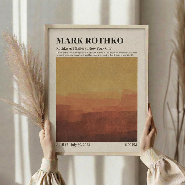 May include: Person holding framed art poster featuring warm gradient bands transitioning from golden yellow at top to deep rust red at bottom. White matted frame with exhibition text for Mark Rothko show at gallery in New York City. Dried wheat stems visible beside frame. Person wears cream colored shirt with gathered cuffs against neutral background.