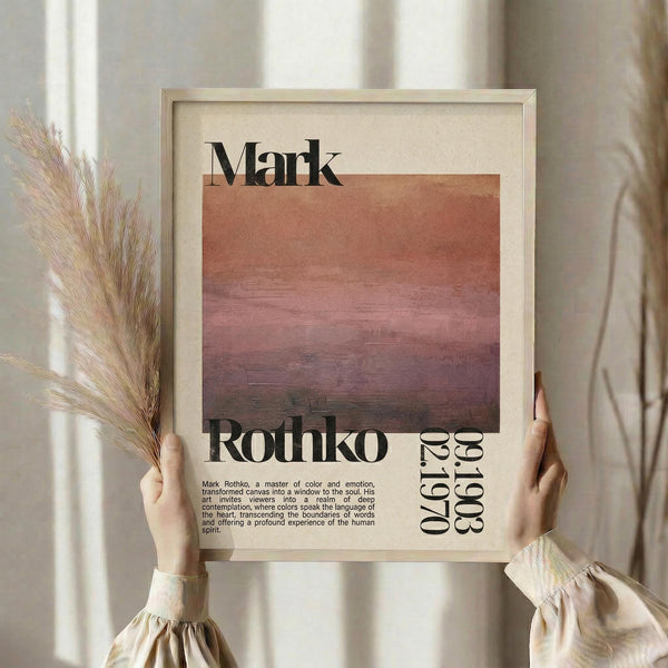 May include: Person holding framed poster print featuring abstract sunset gradient in pink, coral and purple tones with Mark Rothko text and biographical description below. Light wood frame against cream curtained window with dried pampas grass stems visible to left side.