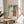 May include: Framed Mark Rothko-style abstract art print displayed on light wood round side table next to beige upholstered chair. Print features vertical composition with blue, white, orange and dark brown color blocks in thin wooden frame. Bright natural light streams across neutral room with white curtains and plant leaves visible.