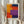 May include: Person holding framed abstract art print with geometric blocks of vibrant orange, red, purple, and navy blue colors in textured brushstroke style. Light wood frame against neutral beige curtained background with dried pampas grass visible on left side.