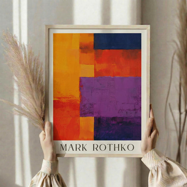 May include: Person holding framed abstract art print with geometric blocks of vibrant orange, red, purple, and navy blue colors in textured brushstroke style. Light wood frame against neutral beige curtained background with dried pampas grass visible on left side.