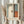 May include: Person holding framed abstract art print featuring soft geometric shapes in muted blue, cream, orange and brown tones with vertical composition. Light wood frame against neutral curtained background with dried pampas grass visible on left side. Soft natural lighting creates gentle shadows.