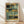 May include: Abstract artwork print in wooden frame leaning against white wall on light wood floor. Painting features geometric blocks and brushstrokes in muted teal, sage green, golden yellow, charcoal black, and cream tones with soft, layered composition. Natural lighting creates diagonal shadows across the textured wall surface.