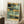 May include: Abstract painting poster with muted teal, sage green, golden yellow, and black geometric shapes and brushstrokes in light wood frame, leaning against white wall corner next to wooden side table with white coffee cup and saucer, beige textured throw blanket visible on white shag rug in bright room with natural window light.