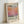 May include: Framed poster print featuring Mark Rothko's name in large black text above a gradient wash of orange, pink, and purple watercolor-like colors. The bottom shows "Rothko" and dates "02.19.03 06.19.69" with small descriptive text. Natural wood frame displayed against white wall, showing dimensional depth and soft lighting shadows.