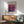 May include: Framed Mark Rothko art exhibition poster with purple and magenta abstract painting, displayed above mid-century modern wooden credenza with white ceramic lamp. Sage green upholstered armchair with wooden frame sits nearby in sunlit room with white walls and warm wood flooring.