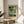 May include: Abstract art print in wooden frame displayed on round wood side table next to beige upholstered chair. Artwork features muted teal, yellow, and black rectangular shapes in painterly style. Natural lighting creates soft shadows on neutral wall. Plant leaves visible in corner of minimalist modern interior setting.