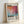 May include: Framed poster featuring Mark Rothko's name in bold black letters above an abstract painting with orange, pink, red, and turquoise color blocks. Bottom shows "Rothko" text with dates "1903-1970" and descriptive paragraph about the artist. Light wood frame against white wall, showing texture and shadows.
