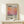 May include: Framed poster print featuring Mark Rothko's name above a gradient square in warm orange, pink and purple tones, with exhibition text below. Displayed on light wood surface with small wrapped gift beside it. Natural lighting creates soft shadows on cream wall background.